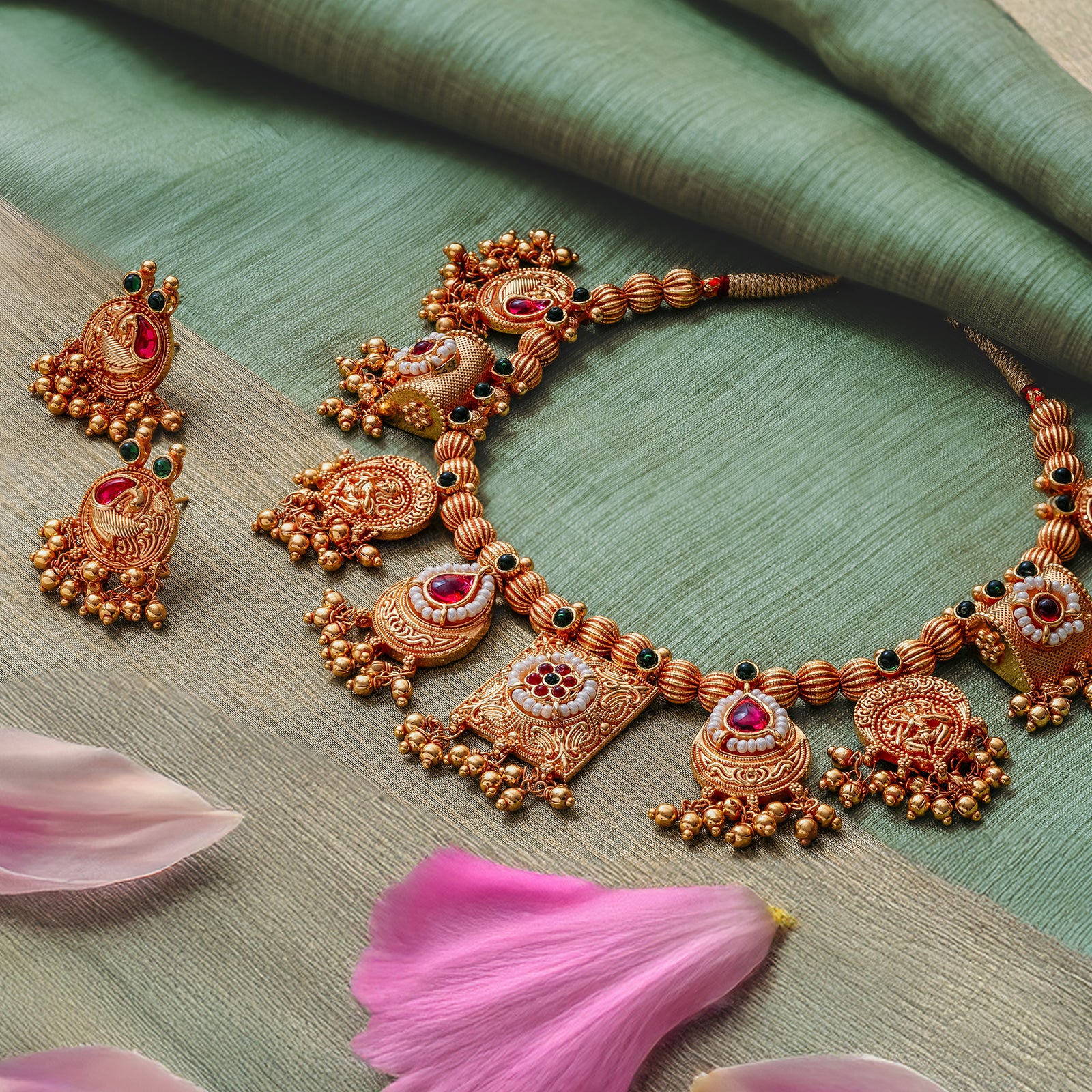 A traditional Indian haar chokda pendant necklace with gold finish and intricate designs, accompanied by matching earrings, displayed on a green fabric with pink petals around.